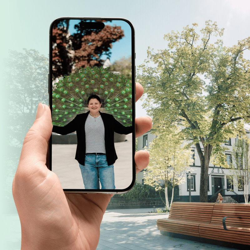 Eine Hand hält ein Smartphone und zeigt ein Bild von einer Frau, die auf dem Marktplatz steht und ein virtuelles Pfauenrad hinter sich hat.