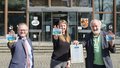 Drei Personen stehen nebeneinander vor der Stadtverwaltung Neuwied. Auf ihren Handinnenflächen, die sie der Kamera entgegenstrecken, kleben Sticker mit dem Fairtrade-Logo. Die Sonne scheint.